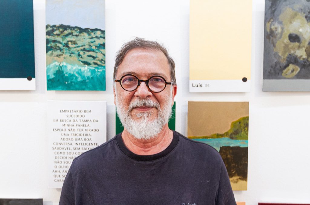 O arquiteto Luiz Amorim, de barba branca e óculos, diante de uma parede com quadros.
