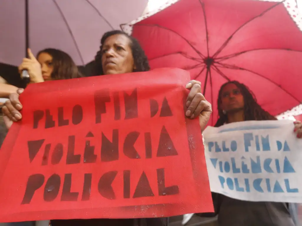 Na foto, um grupo de pessoas participa de uma manifestação sob chuva, protegendo-se com guarda-chuvas enquanto seguram cartazes em português que pedem “pelo fim da violência policial”; os cartazes, em cores como vermelho e branco com letras destacadas, reforçam a mensagem de protesto e solidariedade, transmitindo o clima de mobilização coletiva contra abusos e em defesa dos direitos humanos.