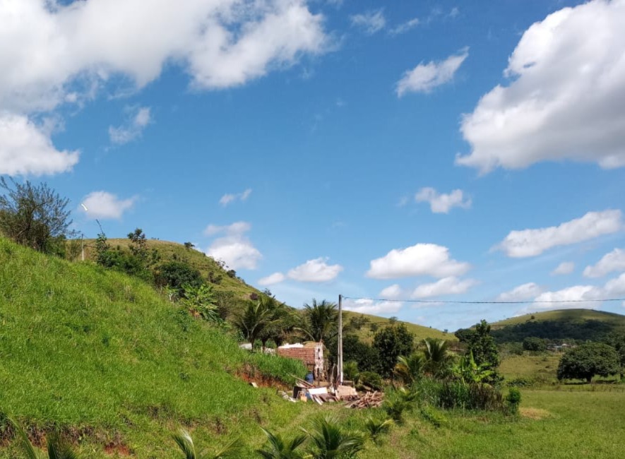 No centro da imagem há uma pequena construção de tijolos, aparentemente simples e parcialmente destruída, cercada por vegetação tropical, incluindo palmeiras e arbustos. Um poste com fios elétricos corta o cenário, indicando presença humana em meio à natureza. O céu azul intenso, pontuado por nuvens brancas e fofas, cria um contraste vibrante com o verde do terreno, transmitindo uma sensação de tranquilidade e amplitude típica de áreas rurais.