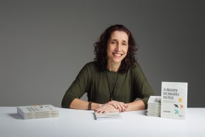 Juliana Romão, mulher branca de cabelos escuros, sorri sentada diante de uma mesa com exemplares do livro A linguagem das mulheres políticas. Veste blusa verde-oliva; fundo cinza neutro