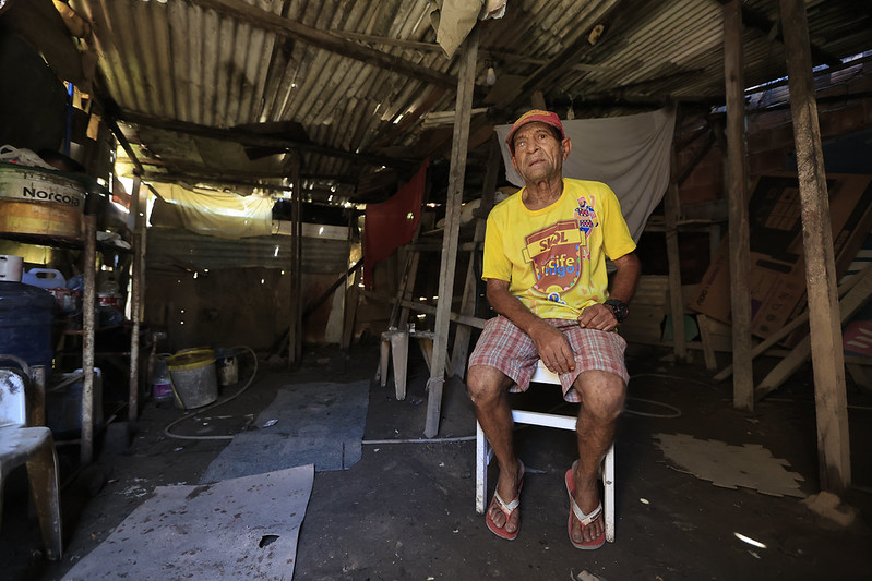 Um homem idoso está sentado em uma cadeira branca no interior de uma casa precária, com piso de terra batida irregular e paredes de madeira cobertas por um telhado de zinco ondulado que deixa entrar raios de luz por entre as frestas. Ele veste camiseta amarela com estampa colorida, shorts xadrez, chinelos vermelhos e um boné listrado, e olha para cima com as mãos sobre as pernas, enquanto ao seu redor se veem baldes, recipientes, um barril com etiqueta, panos pendurados fazendo divisórias e troncos de madeira sustentando a estrutura, num ambiente de pouca luz marcado pelo contraste entre claridades que entram pelas brechas e sombras profundas.
