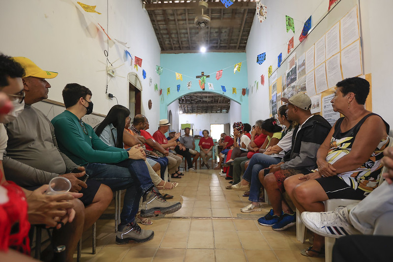 Em uma sala de paredes brancas com piso de cerâmica clara, dezenas de pessoas — homens e mulheres de diferentes idades, alguns de máscara e roupas casuais — estão sentadas em cadeiras plásticas dispostas em duas fileiras que se enfrentam, formando um corredor central, enquanto participam de uma reunião comunitária; o ambiente é decorado com bandeirinhas coloridas de festa junina, fotos e documentos afixados nas paredes, e ao fundo, além de um arco, vê-se uma parede azul-clara com um crucifixo e um ventilador de teto, sugerindo um espaço de caráter religioso ou comunitário.