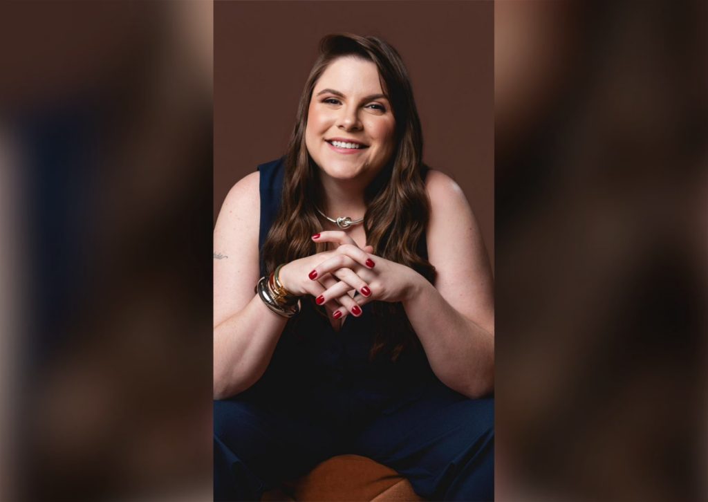 A foto mostra Tabata Tesser sentada contra um fundo marrom, sorrindo de forma calorosa e confiante. Ela tem cabelos longos e castanhos, usa um macacão escuro sem mangas e mantém as mãos entrelaçadas à frente do corpo, exibindo unhas pintadas de vermelho. No pulso, há várias pulseiras metálicas e um relógio; no pescoço, um colar prateado com pingente em forma de nó. A iluminação destaca seu rosto e expressão amigável, transmitindo uma sensação de proximidade e profissionalismo.