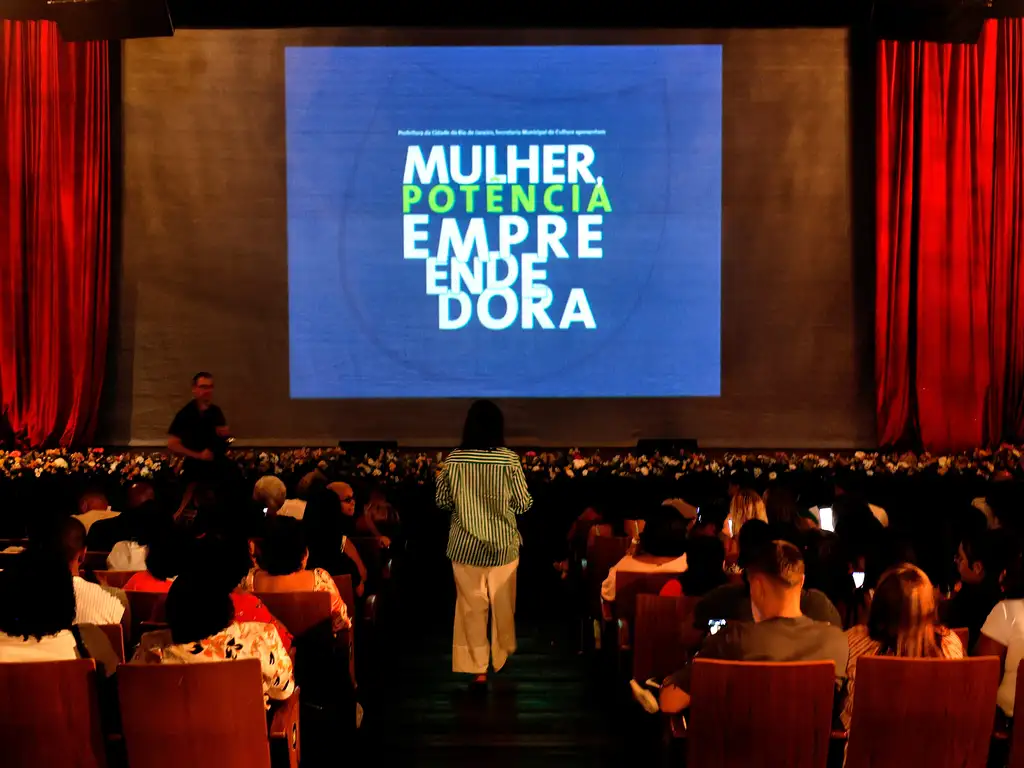 A foto mostra um auditório cheio de pessoas sentadas, voltadas para um palco com cortinas vermelhas abertas e uma grande tela de projeção ao centro. Na tela, lê-se em letras grandes e coloridas “Mulher Potência Empreendedora”, indicando um evento da Prefeitura do Rio de Janeiro voltado ao tema do empreendedorismo feminino. O ambiente é de expectativa e atenção, com luzes baixas e foco na tela, sugerindo o início ou andamento de uma apresentação sobre o poder e a iniciativa das mulheres.