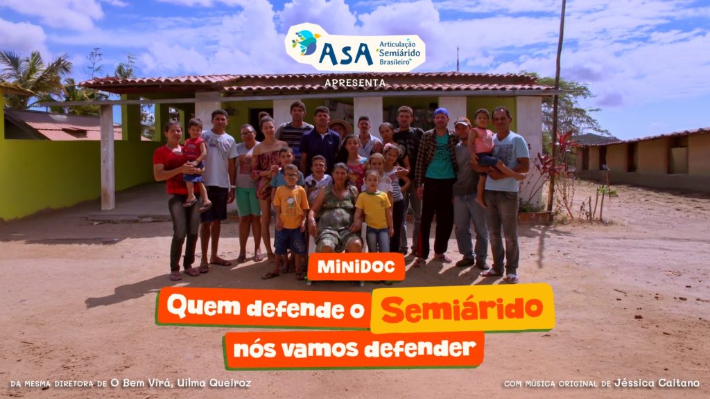 Descrição acessível: Grupo de cerca de vinte pessoas, incluindo adultos, crianças e uma pessoa sentada em uma cadeira, posando em frente a uma construção simples de área rural com telhado de cerâmica e pátio de terra batida sob céu parcialmente nublado. Na parte superior da imagem aparece o logotipo da ASA (Articulação Semiárido Brasileiro) com a palavra “Apresenta”, e no centro há o texto em destaque: “MiniDoc - Quem defende o Semiárido nós vamos defender”; na parte inferior também aparecem créditos de direção e música original.