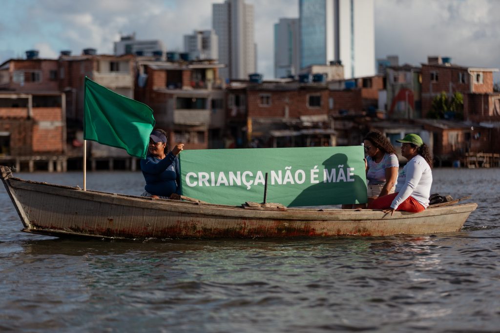 Um barco simples de madeira com três mulheres atravessa o Rio Capibaribe, no Recife, carregando uma bandeira verde e uma faixa com a frase criança não é mãe. Ao fundo, aparecem casas simples e prédios urbanos.