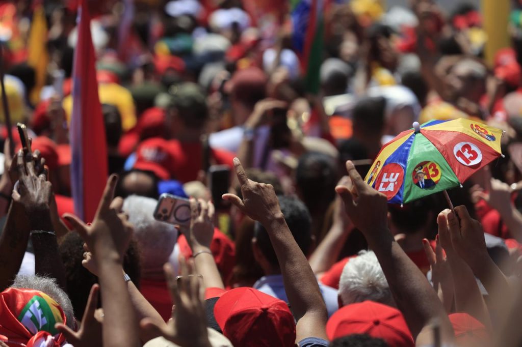 Fotografia de um ato público com grande concentração de pessoas ao ar livre, registrada em plano fechado sobre a multidão. A maior parte dos participantes veste roupas e acessórios vermelhos, enquanto diversos braços erguidos seguram celulares, bandeiras e fazem gestos com as mãos. Em destaque, à direita da imagem, uma pequena sombrinha colorida exibe os números “13” e o nome “Lula” em diferentes painéis. O fundo aparece desfocado, revelando uma massa de pessoas e bandeiras predominantemente vermelhas, amarelas e verdes, o que reforça a dimensão e a densidade do evento. A iluminação forte do sol e as cores vibrantes dos materiais de campanha criam uma cena dinâmica.