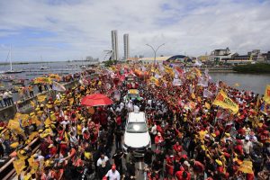 Fotografia aérea de uma manifestação pública em Recife, com grande concentração de pessoas ocupando uma avenida à beira d’água. A multidão carrega bandeiras, faixas e cartazes de apoio ao presidente Lula, enquanto um veículo branco avança pelo centro do ato. Ao lado da via, aparecem o rio, embarcações e uma marina; ao fundo, destacam-se edifícios e outras construções urbanas sob céu parcialmente nublado.