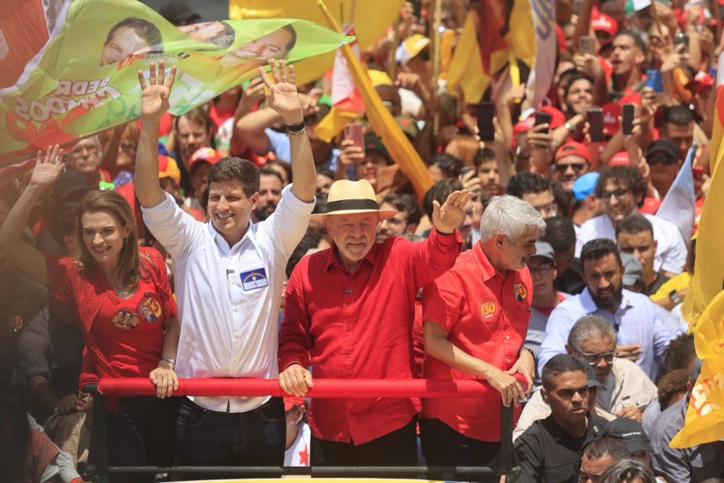 Fotografia de um ato de campanha em via pública, mostrando Lula (de chapéu e camisa vermelha), João Campos (de camisa branca), Marília Arraes e Humberto Costa sobre um veículo ou plataforma cercada por uma grande multidão. As pessoas em destaque estão posicionadas atrás de uma barra vermelha; duas vestem camisas vermelhas, uma veste camisa branca e outra veste roupa vermelha. Algumas acenam para o público enquanto apoiadores ao redor seguram bandeiras e materiais de campanha nas cores vermelha e amarela e registram o momento com celulares. Ao fundo, a multidão ocupa todo o espaço visível, formando uma grande concentração política ao ar livre sob forte luz do dia