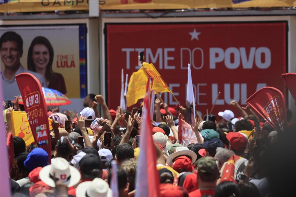 Cena de um ato político ao ar livre, com uma grande multidão reunida em frente a um palanque ou estrutura de campanha. Diversas pessoas ocupam quase toda a imagem, muitas usando bonés e chapéus e levantando os braços, celulares e bandeiras vermelhas, amarelas e brancas. Ao fundo, há grandes painéis de campanha nas cores vermelha e branca, com os textos “TIME DO POVO” e “TIME DE LULA”, além de um cartaz com fotos de duas pessoas e a inscrição “TIME DE LULA”. A iluminação indica um dia ensolarado