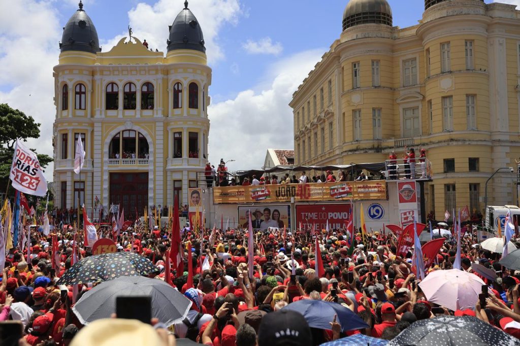 Vista ampla de uma grande manifestação política realizada em uma praça urbana durante o dia, com centenas de pessoas ocupando o espaço em frente a um trio elétrico decorado com faixas e cartazes de campanha nas cores vermelha, branca e amarela. A multidão segura bandeiras, estandartes, celulares e guarda-chuvas para se proteger do sol, enquanto várias pessoas acompanham o evento de uma estrutura elevada e de janelas de edifícios históricos ao fundo. Destacam-se dois prédios de arquitetura clássica em tons amarelo-claro e branco, com torres, cúpulas e grandes janelas, que emolduram a cena. Entre os materiais de campanha, são visíveis palavras como “Time do Povo”, “Time de Lula” e números de candidatos. O céu azul com nuvens brancas, a concentração de bandeiras e o grande número de participantes reforçam a dimensão coletiva e pública do ato.