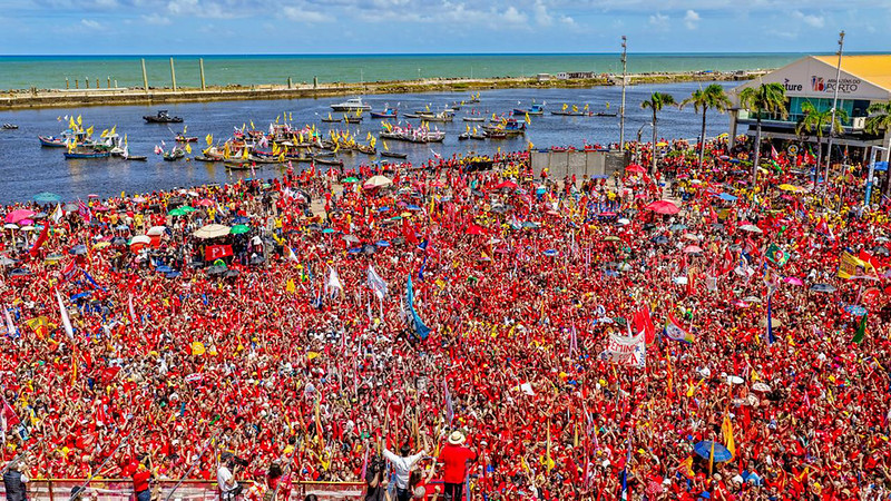 Fotografia panorâmica do Marco Zero, no Recife, mostrando uma grande manifestação pública com milhares de pessoas concentradas na praça e nas áreas próximas ao cais, predominando as cores vermelha e amarela em bandeiras, faixas e cartazes de apoio ao presidente Lula; em primeiro plano, a multidão ocupa praticamente todo o espaço visível, com muitas bandeiras erguidas acima das cabeças, enquanto, ao fundo, aparecem o rio, diversas embarcações decoradas com bandeiras, o quebra-mar e o mar ao horizonte; à direita, vê-se parte da estrutura urbana da área portuária e algumas palmeiras, sob céu parcialmente nublado, compondo uma cena de grande mobilização popular.