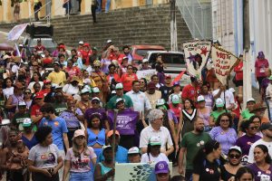 Fotografia de uma caminhada ou manifestação em uma rua histórica. Dezenas de pessoas ocupam a via e descem uma grande escadaria ao fundo. Muitos participantes usam camisetas coloridas, bonés e carregam faixas, cartazes e estandartes com mensagens relacionadas a direitos das mulheres e movimentos sociais. Nas laterais, aparecem prédios antigos em tons claros. A cena é movimentada e mostra uma grande concentração de pessoas reunidas em ato público.