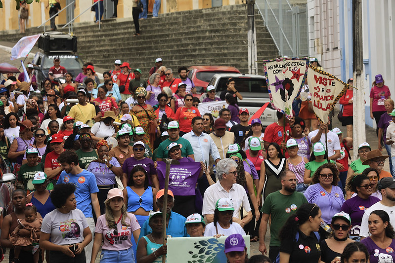 Fotografia de uma caminhada ou manifestação em uma rua histórica. Dezenas de pessoas ocupam a via e descem uma grande escadaria ao fundo. Muitos participantes usam camisetas coloridas, bonés e carregam faixas, cartazes e estandartes com mensagens relacionadas a direitos das mulheres e movimentos sociais. Nas laterais, aparecem prédios antigos em tons claros. A cena é movimentada e mostra uma grande concentração de pessoas reunidas em ato público.