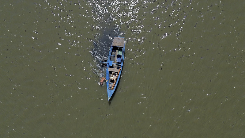 A imagem mostra uma vista aérea de um pequeno barco azul navegando sozinho sobre águas de tom esverdeado. O barco tem formato estreito e alongado, com uma cobertura simples na parte traseira e alguns objetos dentro, como caixas e equipamentos. Um homem aparece próximo ao centro do barco, aparentemente conduzindo ou trabalhando. A luz do sol reflete na superfície da água, criando pontos brilhantes que destacam o contraste entre o movimento do barco e a imensidão tranquila ao redor.