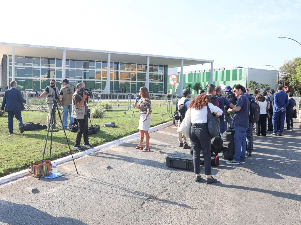 A imagem mostra um grupo de jornalistas e cinegrafistas reunidos em frente a um prédio moderno de arquitetura governamental, com fachada de vidro e colunas brancas. Há câmeras montadas em tripés, microfones e malas de equipamentos espalhados pelo gramado, enquanto uma repórter entrevista alguém. Ao fundo, outras pessoas aguardam em fila próxima a grades de contenção, sugerindo cobertura de um evento oficial ou coletiva de imprensa. O ambiente é claro e organizado, transmitindo o clima típico de uma manhã de trabalho jornalístico.