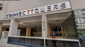 Foto da entrada de um edifício comercial com a placa “CONNECT TOWERS” em destaque na parte superior; a fachada é composta por pilares claros, áreas envidraçadas e uma rampa com corrimãos metálicos no primeiro plano. Próximo à entrada, há uma pessoa apoiada no corrimão utilizando um celular, enquanto outra aparece ao fundo, em um espaço coberto e iluminado. O ambiente tem aparência moderna, com tons neutros predominantes e prédios visíveis ao redor através das superfícies de vidro.