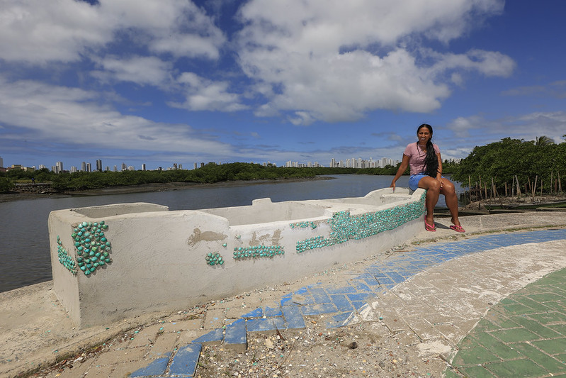 A foto mostra Érika Romão, uma mulher de pele morena, cabelos longos e lisos, sentada sobre uma estrutura de concreto decorada com conchas verdes e azuis, à beira de um rio. Ela veste uma blusa rosa, shorts jeans e tênis vermelhos, sorrindo suavemente enquanto olha para a câmera. O cenário ao fundo revela uma paisagem natural com manguezais e, ao longe, uma linha de prédios sob um céu parcialmente nublado e iluminado pelo sol.