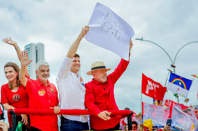 A imagem mostra um ato de campanha política realizado em uma via urbana, com quatro pessoas em destaque sobre um veículo aberto ou trio elétrico (da esquerda para a direita: Marília Arraes, Humberto Costa, João Campos e Lula) -, posicionadas atrás de uma barra de proteção vermelha; uma das pessoas ergue um cartaz branco com uma mensagem escrita à mão, enquanto as demais acenam para a multidão. Todas usam roupas em tons de vermelho e branco, cores também predominantes nas bandeiras e materiais de campanha visíveis ao fundo. Entre os elementos da cena destacam-se uma bandeira vermelha com a sigla “PT”, bandeiras com referências ao estado de Pernambuco, diversos cartazes carregados por apoiadores, postes de iluminação pública e edifícios altos ao longe. O céu aparece encoberto por nuvens, criando uma iluminação suave e difusa.