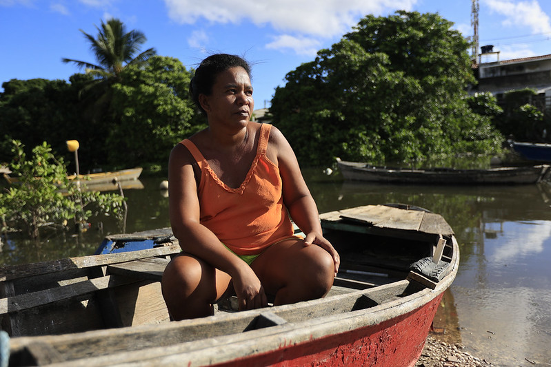 Juliana dos Santos é uma mulher negra sentada no interior de um barco de madeira com casco vermelho, ancorado à beira de um rio de águas escuras e calmas. Ela veste uma blusa regata laranja com renda no decote e short verde, com os braços apoiados sobre as pernas e o olhar sereno voltado para o lado esquerdo da imagem. Ao fundo, a paisagem é composta por vegetação de manguezal densa, coqueiros, outros barcos de madeira parcialmente visíveis e uma pequena construção de concreto sob um céu azul claro com nuvens dispersas, sugerindo uma comunidade ribeirinha em dia ensolarado.