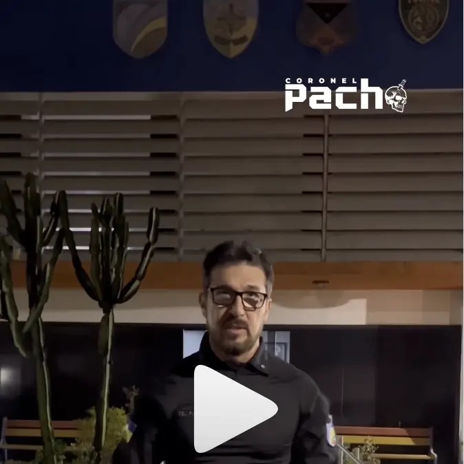 A imagem mostra um homem de óculos e barba, vestindo uma roupa escura com aparência de uniforme, em pé diante de um prédio com painéis horizontais e escudos militares na parede superior. À esquerda, há um cacto alto e, no canto superior direito, aparece o logotipo “Coronel Pach” acompanhado de um pequeno desenho de caveira com faca. No centro, há um ícone de reprodução de vídeo, indicando que se trata de uma captura de um vídeo. A cena parece ter sido gravada à noite ou em ambiente com pouca luz.