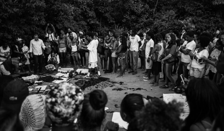 A foto em preto e branco mostra um grupo grande de pessoas reunidas ao ar livre, formando um círculo em torno de um homem que parece conduzir uma fala ou ritual. No centro, há objetos dispostos no chão — tecidos, flores e papéis — enquanto os demais observam atentamente. A cena transmite um momento de coletividade e expressão comunitária, ligado a uma homenagem, protesto ou cerimônia simbólica, em meio à vegetação densa ao redor.