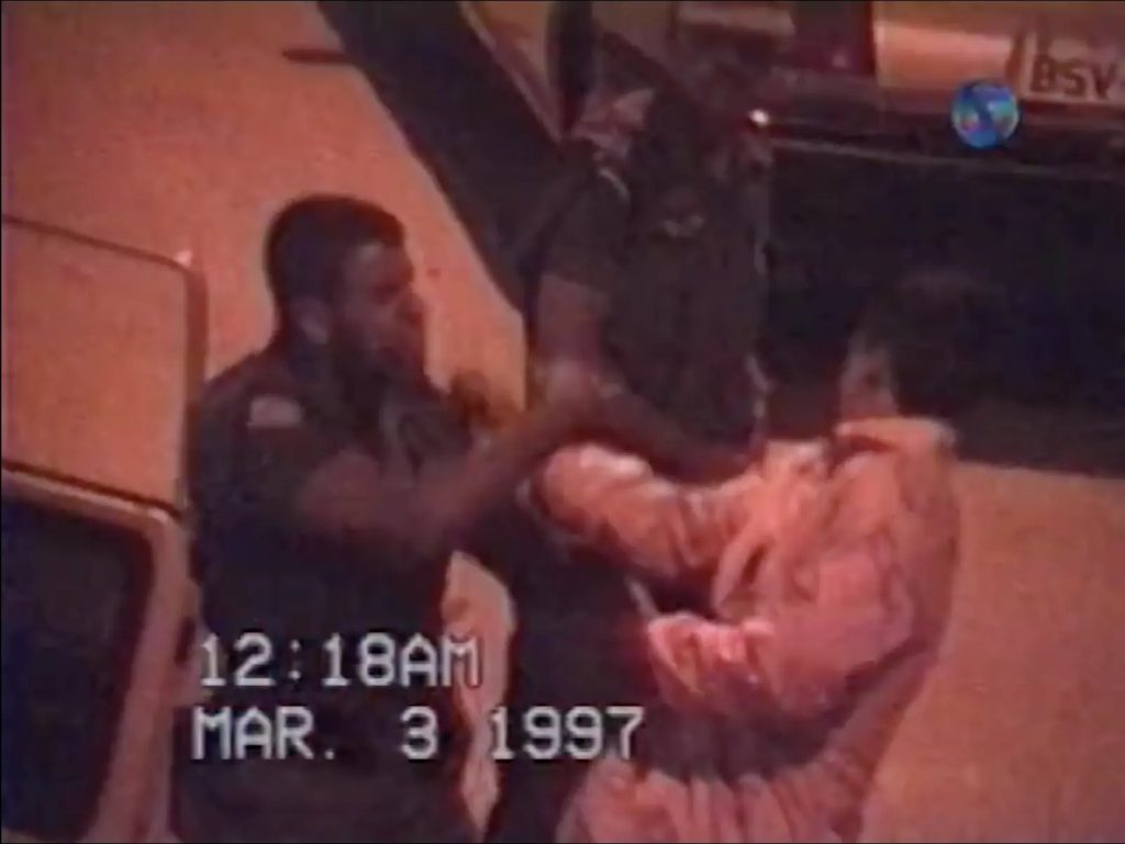 A imagem parece ser um registro de vídeo datado de 3 de março de 1997, às 12h18 da madrugada, mostrando dois policiais uniformizados interagindo de forma tensa com uma pessoa vestida com um casaco claro, em uma rua iluminada artificialmente. O enquadramento e a qualidade granulada sugerem uma gravação de câmera de segurança ou reportagem televisiva. Ao fundo, há um veículo estacionado, e o ambiente transmite uma atmosfera de abordagem policial noturna.