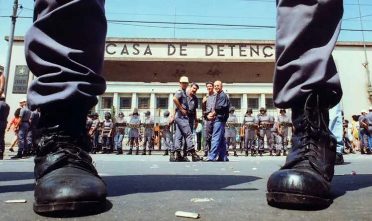 A imagem mostra a fachada da Casa de Detenção, com um grupo de policiais fardados posicionados em frente ao prédio. A foto é tirada de um ângulo baixo, destacando as botas pretas de dois policiais em primeiro plano, o que transmite uma sensação de autoridade e tensão. Ao fundo, há uma fileira de agentes com escudos e capacetes, formando uma barreira de segurança, enquanto alguns homens conversam próximos à entrada do edifício.