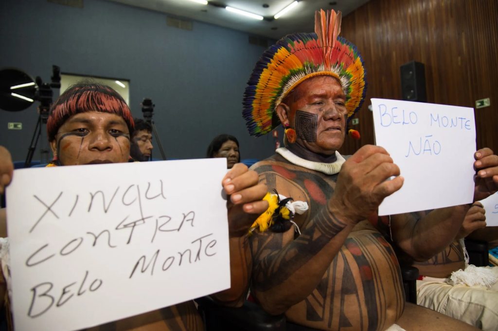 A foto mostra dois homens indígenas sentados em um ambiente interno, segurando cartazes escritos à mão com mensagens de protesto contra a usina de Belo Monte. Ambos estão com pinturas corporais tradicionais e adornos coloridos, incluindo cocares de penas vibrantes. Ao fundo, outras pessoas e câmeras indicam que o momento ocorre em um espaço público, possivelmente uma audiência ou reunião.