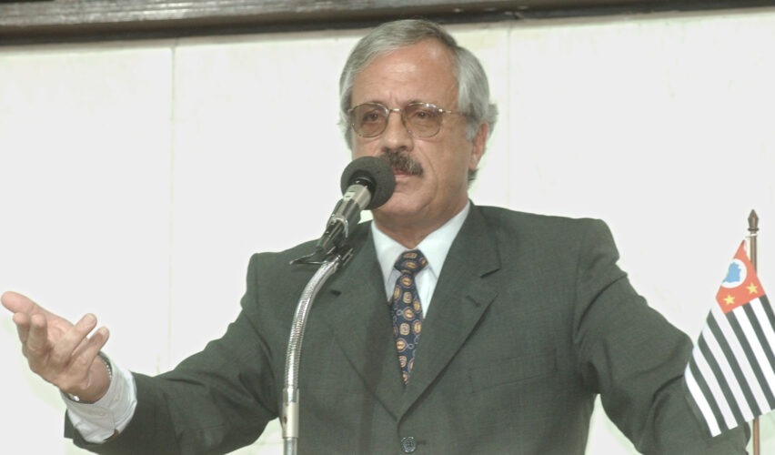 A foto mostra Ubiratan Guimarães, político e ex-coronel da Polícia Militar de São Paulo, falando em um púlpito com microfone. Ele veste terno escuro, camisa branca e gravata estampada, e aparece gesticulando com uma das mãos enquanto discursa. Ao lado do microfone há uma pequena bandeira do estado de São Paulo, reforçando o contexto institucional da cena. O ambiente é formal, com fundo claro e iluminação uniforme, típico de uma sessão ou pronunciamento oficial.