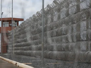 A imagem mostra uma cerca de segurança composta por várias camadas de arame farpado enrolado, formando uma barreira densa e impenetrável ao longo de um muro. Ao fundo, há uma torre de vigilância marrom com janelas e câmeras, típica de instalações de alta segurança como prisões ou áreas militares. O chão é coberto por cascalho escuro e o céu está nublado, criando uma atmosfera de isolamento e controle.