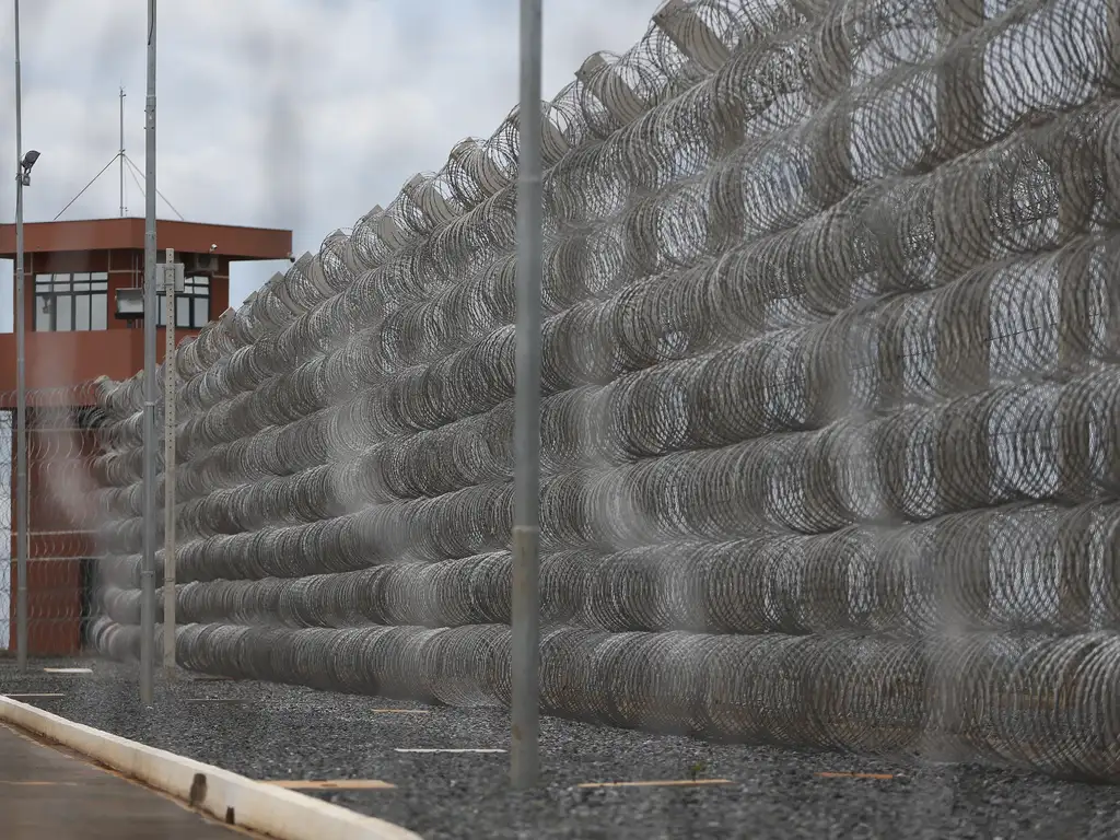 A imagem mostra uma cerca de segurança composta por várias camadas de arame farpado enrolado, formando uma barreira densa e impenetrável ao longo de um muro. Ao fundo, há uma torre de vigilância marrom com janelas e câmeras, típica de instalações de alta segurança como prisões ou áreas militares. O chão é coberto por cascalho escuro e o céu está nublado, criando uma atmosfera de isolamento e controle.