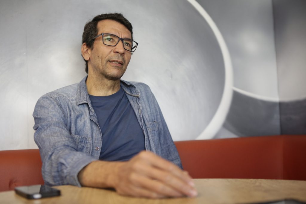 Humberto Barbosa está sentado à mesa em um ambiente interno, usando óculos, camiseta azul e camisa jeans aberta; tem cabelos curtos e escuros e aparece em posição de conversa, com uma das mãos apoiada sobre a mesa. Ao fundo, há uma parede cinza com formas circulares claras e um banco estofado vermelho, compondo uma cena de entrevista ou reunião.