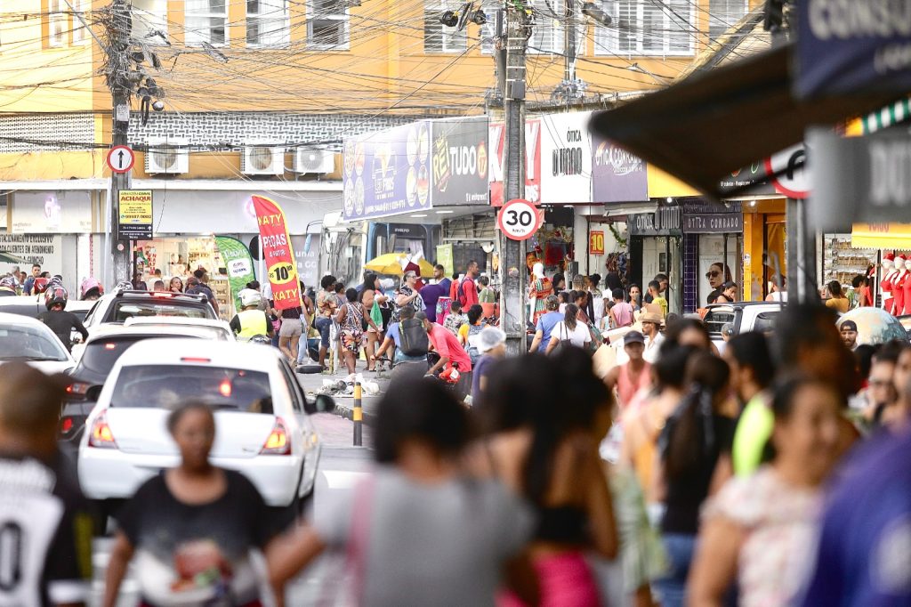 Rua comercial movimentada em área urbana, com grande circulação de pedestres nas calçadas e veículos trafegando lentamente pela via; ao longo da rua há diversas lojas com placas e vitrines, além de postes carregados por muitos fios elétricos e de telecomunicações suspensos sobre a via, compondo uma cena de intenso movimento cotidiano, comércio ativo e alta densidade de infraestrutura urbana.