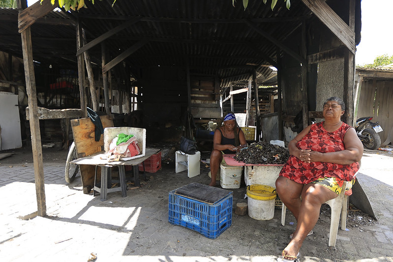 A imagem mostra duas mulheres em um espaço simples e rústico, coberto por telhas de zinco e sustentado por madeiras. À direita, uma mulher de pele escura, corpo robusto e cabelos curtos está sentada em uma cadeira plástica branca, vestindo uma blusa vermelha estampada e shorts coloridos, com expressão serena e olhar voltado para cima. À esquerda, mais ao fundo, outra mulher de pele escura e porte magro usa uma bandana azul e roupas escuras, concentrada em separar ou limpar conchas sobre uma mesa improvisada. O ambiente é modesto, com caixas plásticas, baldes e mesas simples, sugerindo um local de trabalho artesanal ou comunitário.