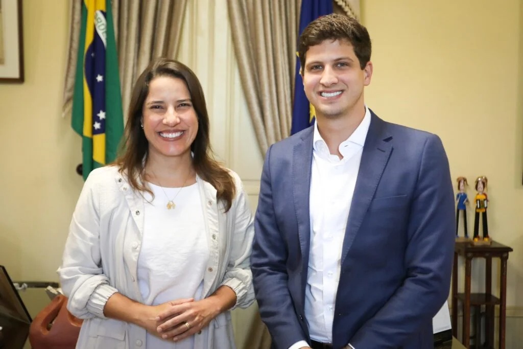 A foto mostra Raquel Lyra e João Campos sorrindo lado a lado em um ambiente formal. Raquel está à esquerda, vestindo uma blusa branca e um casaco claro; João está à direita, com terno azul e camisa branca. Ao fundo, aparecem as bandeiras do Brasil e de Pernambuco, além de cortinas bege e uma mesa com pequenas figuras decorativas. A imagem transmite um clima de cordialidade e profissionalismo, típico de um encontro institucional.