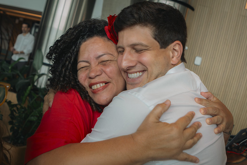 Dani Portela e João Campos aparecem em primeiro plano, abraçadas de forma próxima. Dani Portela é uma mulher negra, veste roupa vermelha e usa um acessório vermelho no cabelo; João Branco é um homem branco, jovem e veste camisa branca. Os rostos estão encostados e ambos estão com os olhos fechados durante o abraço. A foto foi feita em ambiente interno, com alguns vasos de plantas e outras pessoas desfocadas ao fundo. A iluminação é suave e destaca o gesto de proximidade entre as duas pessoas.
