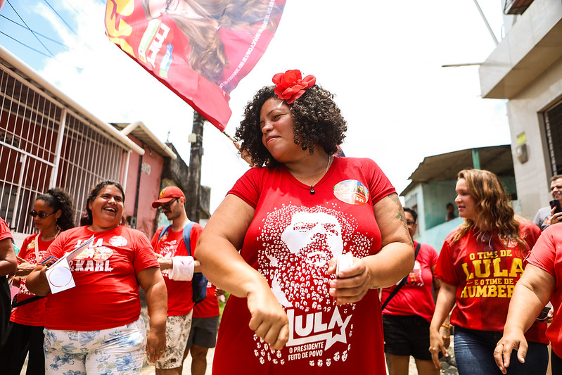 Dani Portela, uma mulher negra, de cabelo preto cacheado, usando uma camiseta vermelha com estampa e textos políticos, caminha por uma rua acompanhada por um grupo de pessoas que também vestem roupas vermelhas. Ao fundo, há uma bandeira vermelha erguida e casas dos dois lados da via. A cena acontece durante o dia, em ambiente externo, e mostra uma atividade coletiva em espaço urbano.
