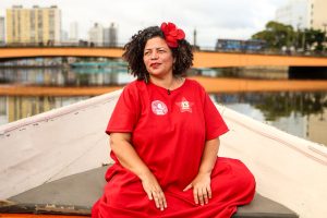 Dani Portela, mulher negra com cabelo preto cacheado e adorno vermelho na cabeça está sentada na proa de um barco, vestindo roupa vermelha. Ao fundo, aparecem um rio de águas calmas, uma ponte alaranjada e edifícios urbanos. A cena ocorre durante o dia, em ambiente externo, com a cidade refletida na água.