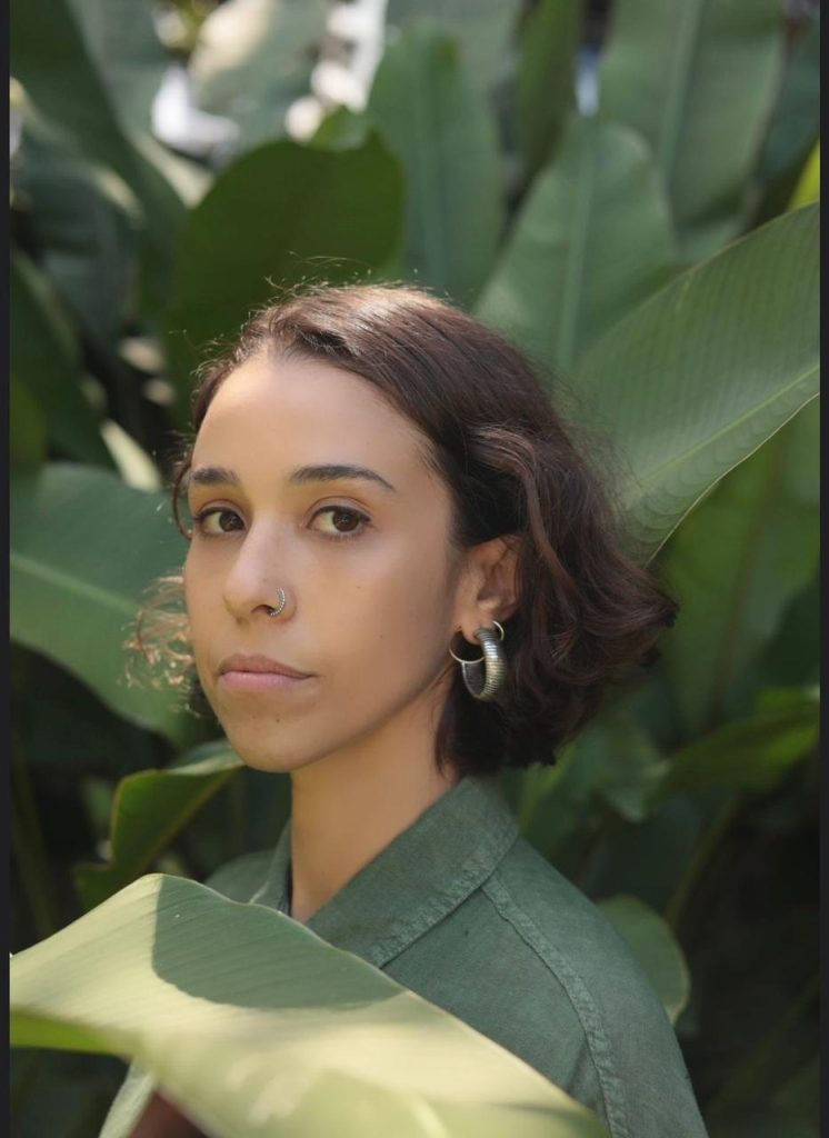 Retrato de Ana Carolina Marsicano, mulher jovem de cabelos castanhos curtos, em meio a grandes folhas verdes de vegetação tropical. A pessoa está voltada para a câmera, com cabelos curtos escuros, usando camisa verde, argola no nariz e brincos metálicos grandes. A fotografia tem iluminação natural suave e fundo desfocado, destacando o rosto entre as folhas.
