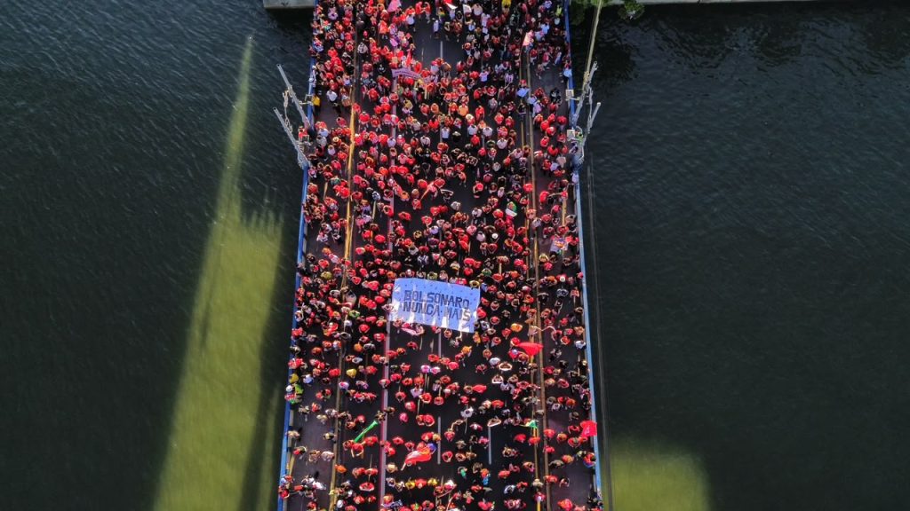 Fotografia aérea mostra uma multidão ocupando uma ponte sobre o rio Capibaribe. Milhares de pessoas estão reunidas, muitas vestidas de vermelho e usando bonés da mesma cor. No centro da manifestação, um grupo segura uma faixa branca com letras azuis, onde se lê “Bolsonaro Nunca Mais”. Entre os manifestantes, aparecem bandeiras e outros materiais vermelhos. A ponte atravessa verticalmente a imagem, de uma extremidade à outra, cercada pelas águas escuras do rio. A luz do sol ilumina trechos da água, formando grandes manchas esverdeadas que contrastam com o tom escuro do rio e o vermelho predominante na manifestação.
