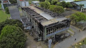 Fotografia aérea mostra um prédio de laboratórios da Universidade Federal de Pernambuco (UFPE) após um incêndio. O edifício de vários andares apresenta danos severos, com parte do telhado destruída, estruturas metálicas retorcidas, janelas quebradas e paredes enegrecidas pelo fogo. Escombros estão espalhados ao redor da construção. O prédio é cercado por árvores de copas verdes e por outras edificações aparentemente preservadas. Duas pessoas aparecem caminhando pela via ao lado do edifício, evidenciando a dimensão da estrutura atingida.