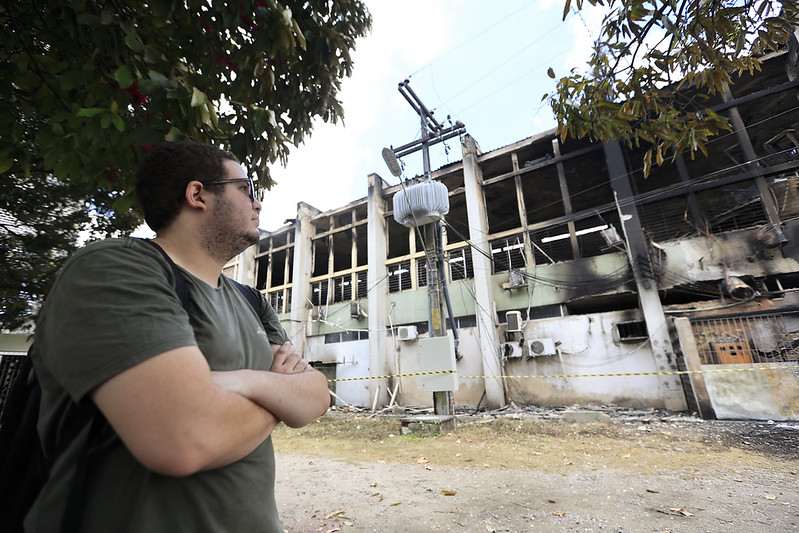 Na imagem, jovem estudante, com braços cruzados e olhar distante, observa o que restou do prédio após o incêndio da UFPE. O prédio, de dois pavimentos, parcialmente destruído, mostra sinais do fogo