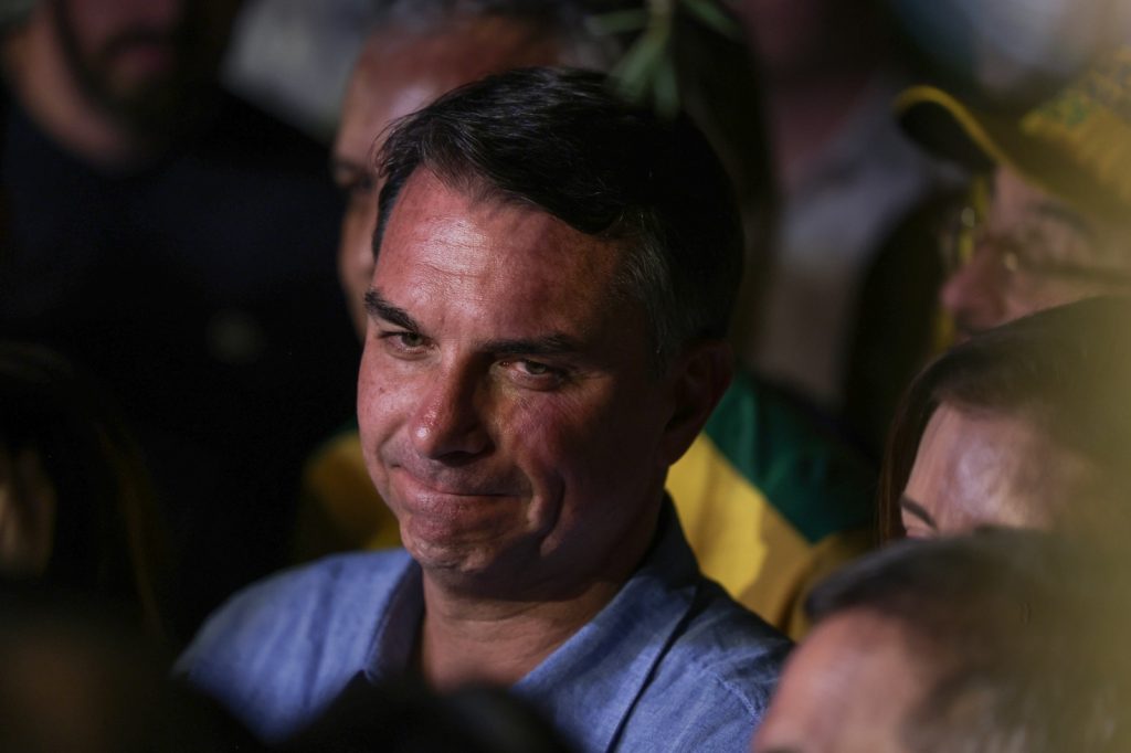 Foto de Flávio Bolsonaro, homem branco de cabelos curtos, em ambiente escuro, cercado de outras pessoas das quais só aparecem os vultos e sombras.