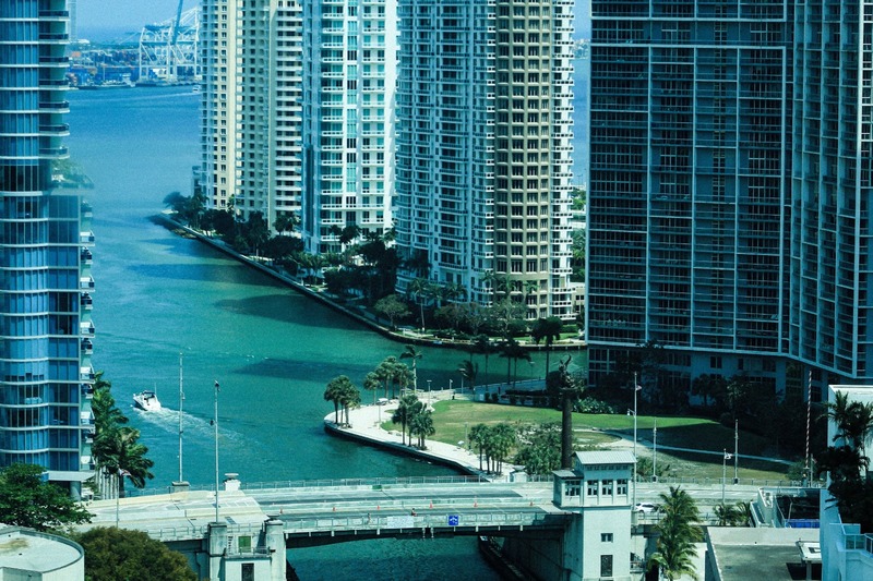 A imagem retrata uma vista urbana característica de Miami, com modernos arranha-céus à beira de um canal de águas azul-esverdeadas, onde uma pequena embarcação navega tranquilamente. A cena combina a arquitetura vertical da cidade com áreas arborizadas de palmeiras e uma ponte que cruza o canal, transmitindo a atmosfera sofisticada, vibrante e costeira que marca a paisagem de Miami.