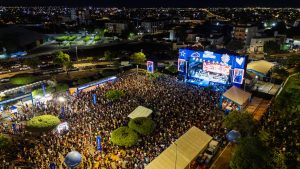Vista aérea noturna de um grande evento ao ar livre realizado em área urbana, mostrando uma multidão muito numerosa concentrada diante de um palco iluminado por luzes azuis e telões laterais. Ao redor do público há tendas, estruturas de apoio, banheiros químicos, árvores e vias com veículos estacionados ou em circulação. A cidade se estende ao fundo sob o céu escuro, com milhares de pontos de luz provenientes de ruas, prédios e residências, criando um amplo panorama urbano. A iluminação intensa do palco contrasta com as áreas mais escuras ao redor, destacando a dimensão do público e a atmosfera festiva do evento.