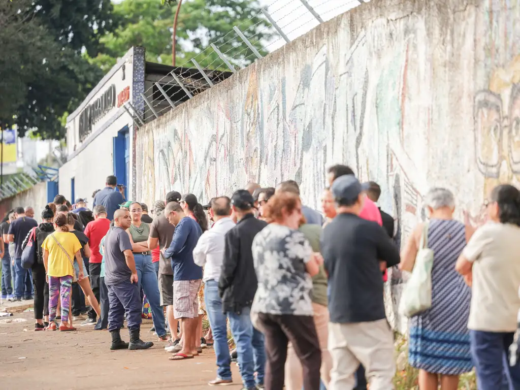 Fila de pessoas em uma calçada ao lado de um longo muro claro com grafites desgastados. As pessoas, vestindo roupas casuais e carregando bolsas ou mochilas, formam uma fila extensa que segue ao longo do muro. Ao fundo, há árvores com folhagem verde e uma construção de fachada branca e azul. A cena acontece ao ar livre, durante o dia, e transmite a ideia de espera ou atendimento em um local movimentado.