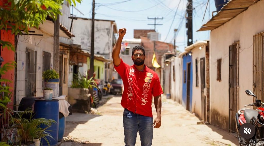 Jones Manoel está posicionado no centro de uma rua estreita de bairro residencial, com o braço direito levantado e o punho fechado acima da cabeça. Ele é um homem negro, de barba e óculos, e está usando uma camisa vermelha com estampas claras na frente, calça jeans e óculos. A rua é de chão claro e é cercada por casas simples, próximas umas das outras, com portas, janelas gradeadas e fachadas em tons variados de branco, azul, verde e outras cores suaves. Há postes e fios elétricos ao longo da via, algumas plantas e vasos nas calçadas, um tambor azul à esquerda e uma motocicleta parcialmente visível à direita. A cena está iluminada por luz solar intensa, criando sombras marcadas e destacando a profundidade da rua ao fundo.