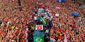 A imagem mostra uma grande manifestação política em uma avenida lotada, com milhares de pessoas reunidas ao redor de um veículo de campanha onde está o presidente Lula segurando uma bandeira do Brasil. Diversas bandeiras, cartazes e faixas nas cores vermelha, verde e amarela são visíveis entre a multidão, incluindo mensagens de apoio e o número “13” estampado em materiais de campanha. A cena é capturada de um ponto elevado, destacando a densidade do público e o caráter coletivo do evento.
