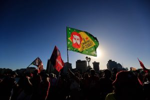 Fotografia de uma manifestação ao ar livre no final da tarde, com várias pessoas reunidas em primeiro plano, vistas principalmente em silhueta contra a luz do sol. Ao centro, destaca-se uma grande bandeira verde com a frase “Brasil é nosso” e a ilustração estilizada de uma figura humana, enquanto outras bandeiras vermelhas são erguidas ao redor. Ao fundo, aparecem edifícios urbanos e postes de iluminação sob um céu azul limpo, criando forte contraste entre a luminosidade do horizonte e as pessoas na cena.